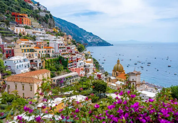 Colorful buildings cascade down a cliffside along the Amalfi road, overlooking a sparkling blue sea dotted with boats.