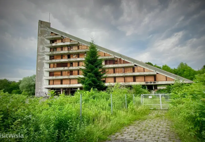 Opuszczony budynek o trójkątnym kształcie, z oknami zabitymi deskami, otoczony bujną zielenią. Idealne urbex miejsca dla poszukiwaczy przygód.