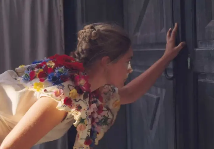Una ragazza con un vestito fiorito, capelli raccolti, spinge una porta scura. Un'immagine che evoca la recensione di Midsommar.