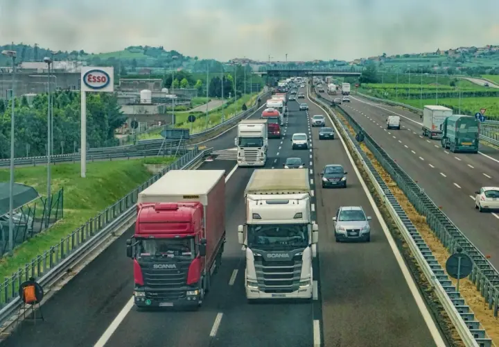Ciężarówki na autostradzie, jedne wymijają, inne omijają. W tle znak Esso i zielone wzgórza.