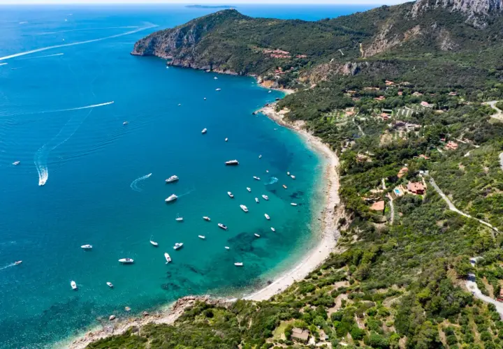 Baia con acque cristalline e numerose barche, incastonata tra la costa verdeggiante e le pendici del monte Argenia.