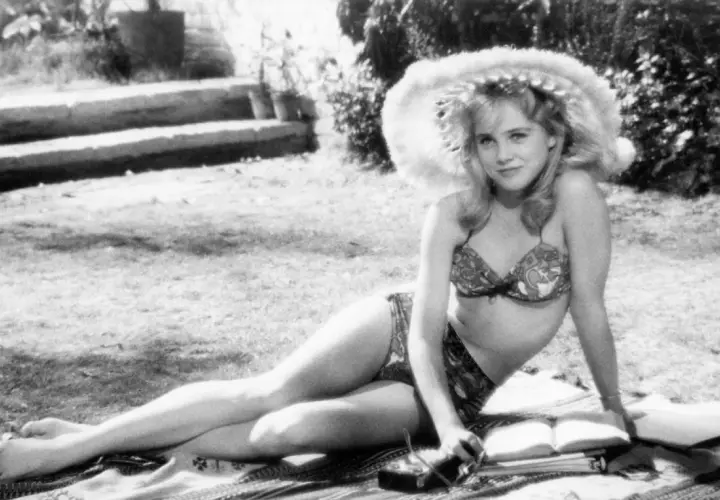 Młoda kobieta w bikini i kapeluszu leży na trawie, czytając książkę. Scena jak z letnich filmów erotycznych.