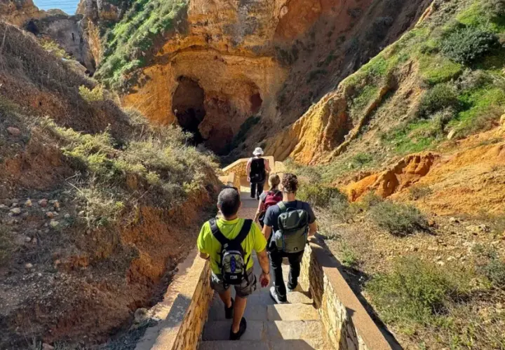 Hikers descend a stone staircase on a scenic trail in Portugal, leading towards a large cave entrance nestled within rugged, sun-drenched cliffs.