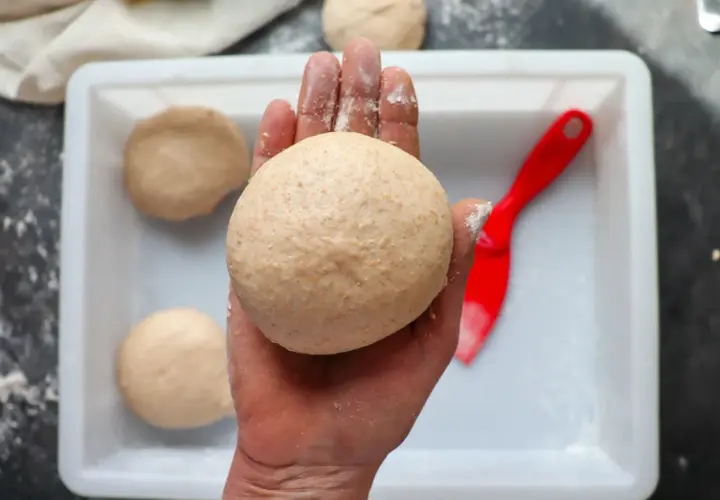Hand hält klebrigen Pizzateig. Mehr Teiglinge in einer weißen Schale, daneben ein roter Teigschaber.