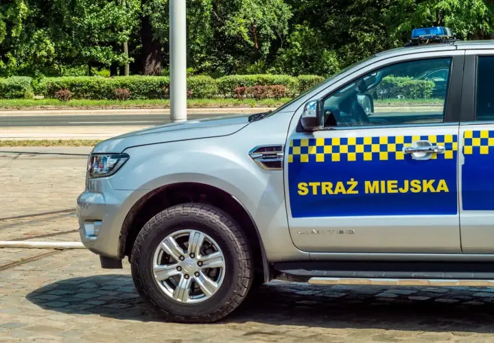 Srebrny samochód Straży Miejskiej z niebiesko-żółtym pasem i napisem "STRAŻ MIEJSKA".