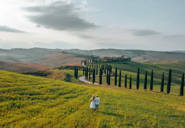 A couple enjoys the view of rolling hills and a cypress-lined road, perfect for renting a car in Tuscany.
