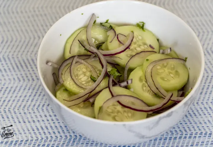 Deliciosa ensalada de pepino con cebolla morada y hierbas frescas, servida en un tazón blanco sobre un mantel azul y blanco.