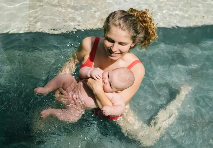 Mamma sorridente tiene in braccio il suo neonato durante un corso di nuoto per neonati in piscina.