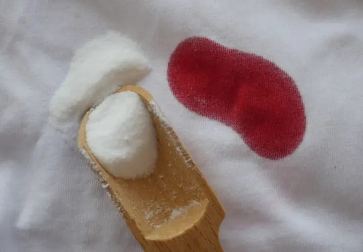 Une cuillère en bois contient une poudre blanche à côté d'une tache rouge sur un tissu blanc. Une tache de sang qui ne part pas au lavage, c'est le cauchemar de tout parent.