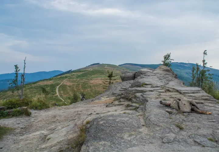 Kamienista ścieżka prowadzi na beskid slaski szczyt. W oddali widać zielone wzgórza i niebieskie pasma górskie pod zachmurzonym niebem.