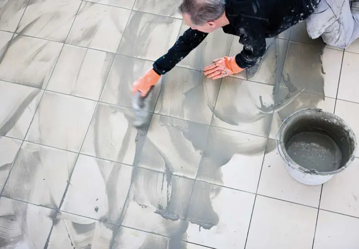 Man med orange handskar arbetar med att måla fogar på ett kaklat golv. En hink med fogmassa står bredvid.