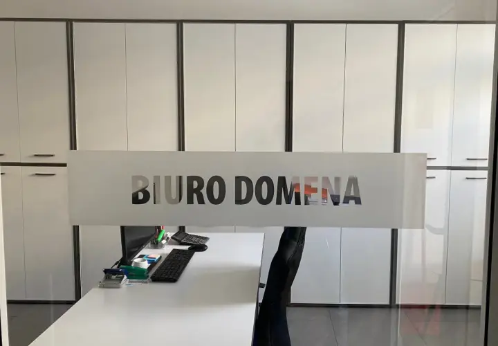 Szklana witryna z napisem "BIURO DOMENA" na tle nowoczesnego biura z białymi szafami.