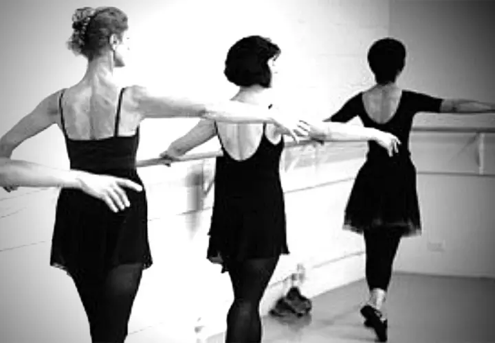 Three women in black leotards practice ballet at a barre.