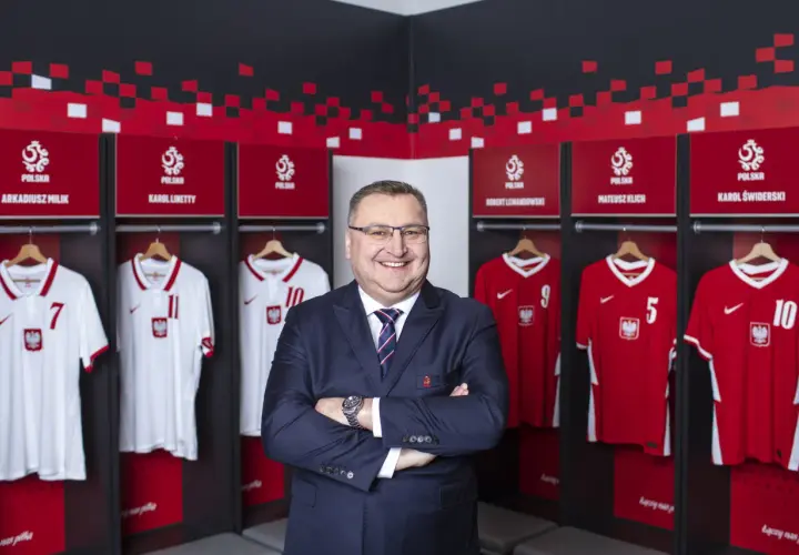 Uśmiechnięty mężczyzna w garniturze stoi przed szatnią reprezentacji Polski. Widać tam koszulki z nazwiskami znanych piłkarzy, tworząc zgrupowanie reprezentacji Polski.
