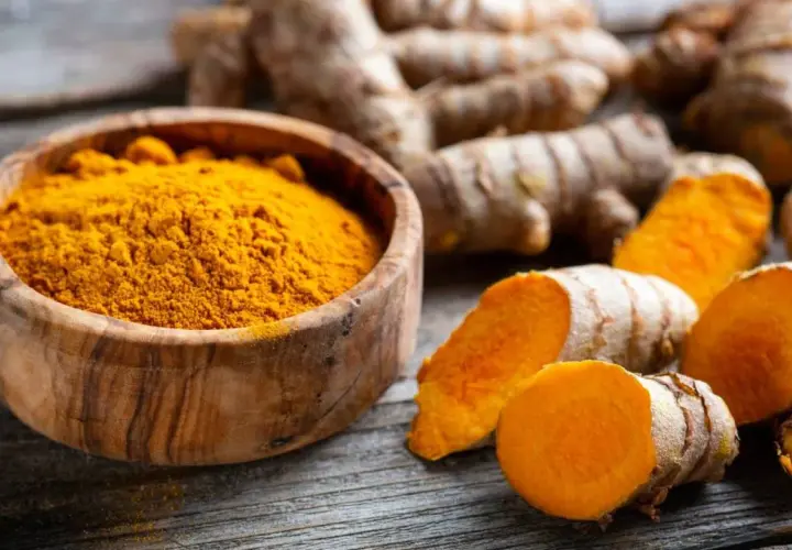 Raíces de cúrcuma y polvo dorado en cuenco de madera, evocando las propiedades de la cúrcuma en cápsulas.
