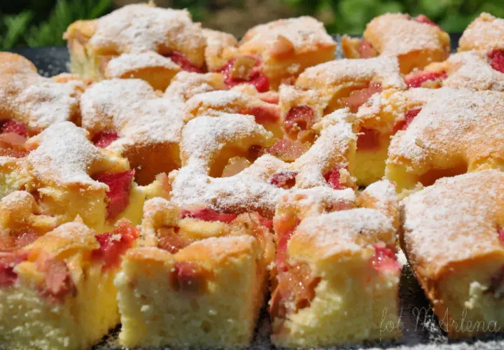 Szybkie ciasto jogurtowe z rabarbarem, posypane cukrem pudrem. Kawałki ciasta z widocznymi różowymi kawałkami rabarbaru.
