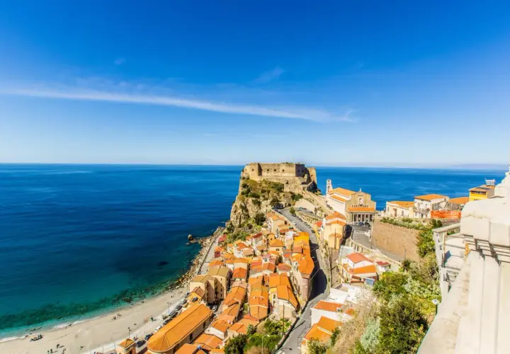 A stunning view of Scilla, Calabria, with its colorful houses cascading down to a beach and the ancient Ruffo Castle perched on a cliff overlooking the turquoise sea. This is what to see in Calabria!