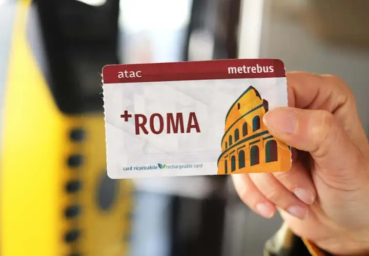Una mano tiene una tessera metropolitana Roma con il Colosseo stilizzato. La carta è ricaricabile e riporta il logo ATAC.
