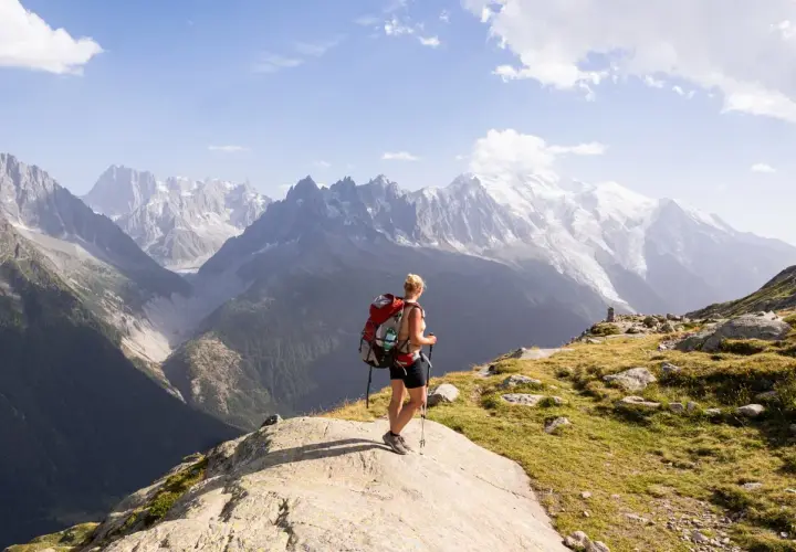 A hiker with a red backpack pauses on a rocky outcrop, enjoying the breathtaking view of the Mont Blanc massif during a challenging hike.