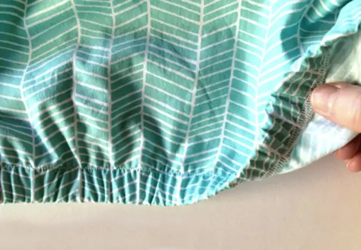 Close-up of a hand demonstrating how to sew in elastic on a teal and white chevron patterned fabric. The elastic creates a gathered, ruffled effect.