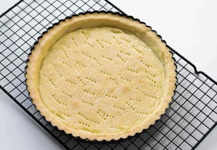Base di pasta sucrée dorata e forata, pronta per essere farcita, su una griglia nera.