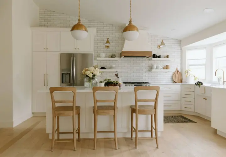 A bright, airy modern farmhouse kitchen with white subway tile, gold pendant lights, and a large island with three wooden stools.