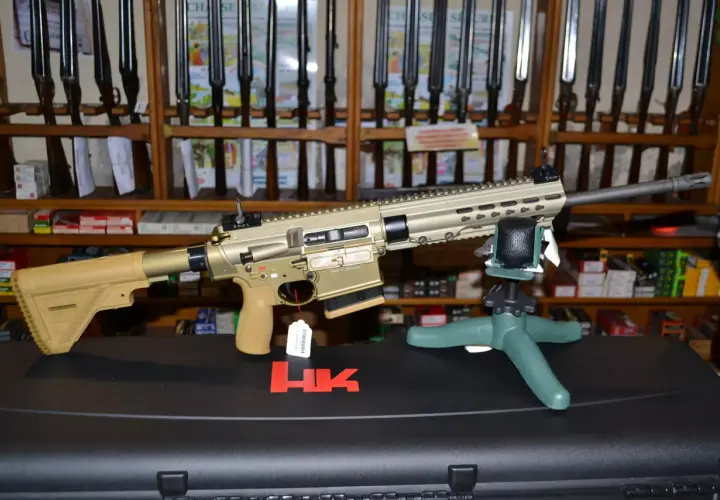 Un fusil HK MR 308, de couleur sable, est présenté sur un support. Derrière, des étagères chargées de fusils et de boîtes de munitions.