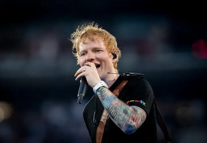 Ed Sheeran śpiewa z mikrofonem, jego ręka ozdobiona kolorowymi tatuażami.