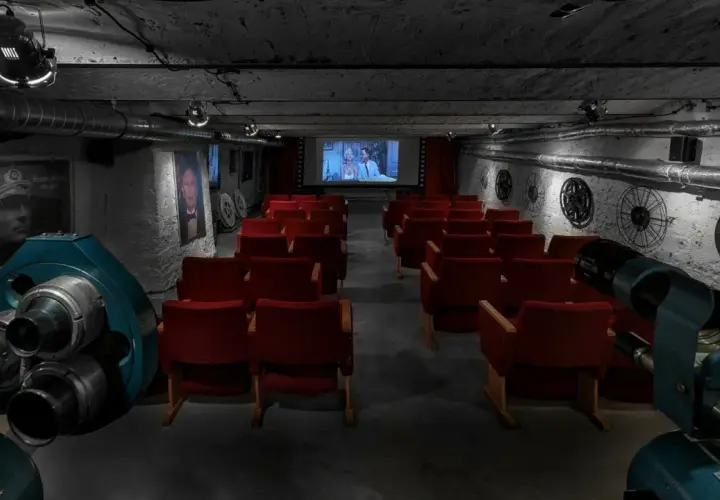 Kameralna sala kinowa z czerwonymi fotelami i zabytkowym projektorem. W tle ekran z filmem i dekoracje nawiązujące do historii kina, jakby to było pierwsze kino w Polsce.