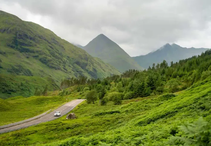 A car drives on a winding road through lush green Scottish Highlands, perfect for a hiking itinerary.