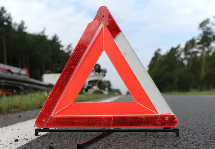 Trójkąt ostrzegawczy, część obowiązkowego wyposażenia samochodu, stoi na poboczu drogi. W tle widać przewrócony samochód ciężarowy.