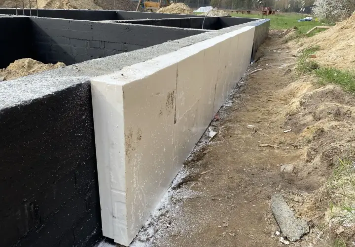 Izolacja fundamentów z białych płyt styropianowych przylega do czarnych bloczków betonowych fundamentowych.