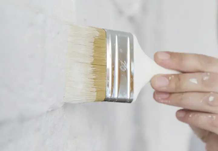 Pennello bianco stende pittura su muro grezzo. Si può mischiare il fissativo nella pittura per una finitura migliore.