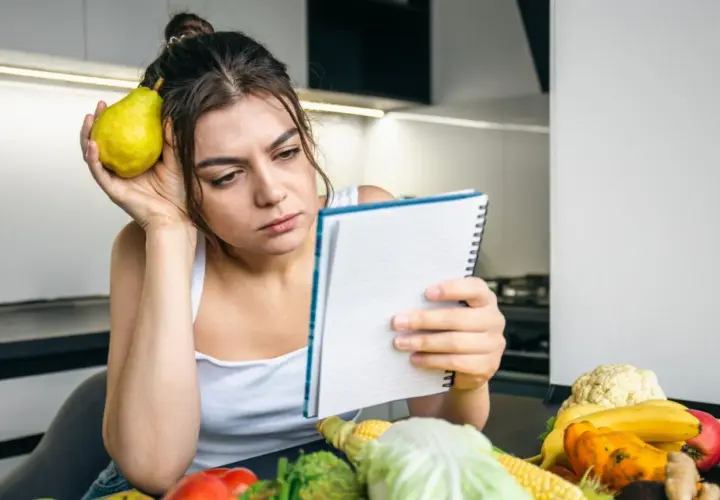 Zamyślona kobieta z gruszką przy uchu przegląda notatnik. Na stole warzywa i owoce, jakby planowała dietę 500 kcal.