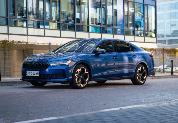 Elegancka, błękitna Skoda Superb 2025 z nowoczesnymi felgami stoi przed szklanym budynkiem.