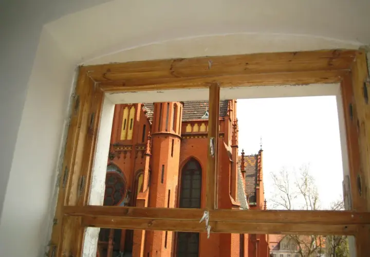 Widok przez stare okna skrzynkowe na ceglaną, zabytkową budowlę z wieżami i ozdobnymi detalami.