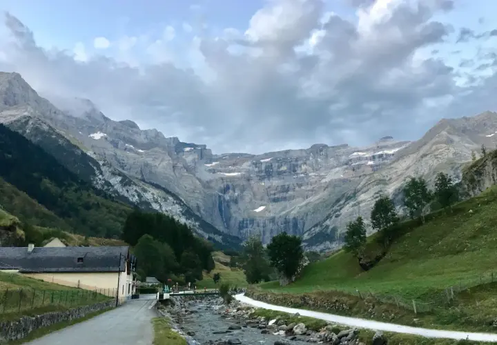 Un camino serpentea junto a un río rocoso hacia el imponente pico Taillón, con una cascada visible en la distancia y un pueblo a la izquierda.