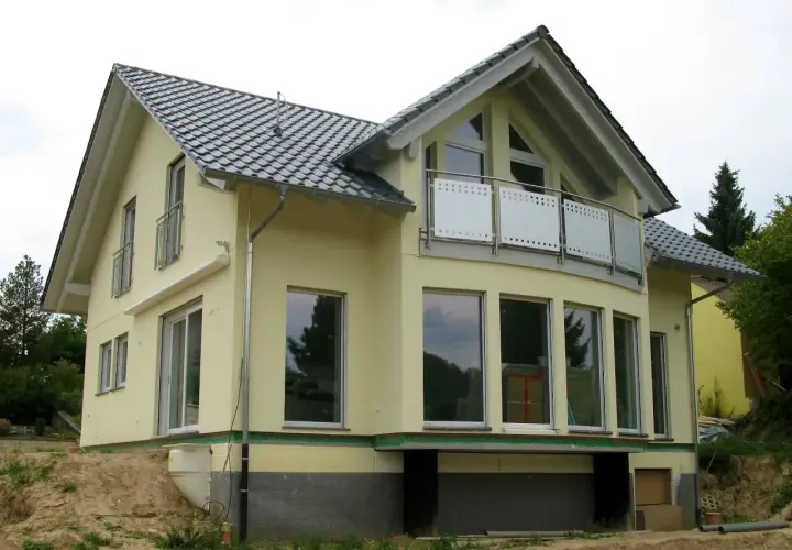 Nowy dom z żółtą elewacją i dachem w kształcie litery L. Duże okna i balkon dodają mu nowoczesnego charakteru.