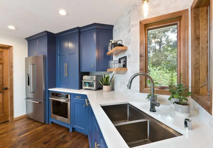 Stylish kitchen sink styling with a modern faucet, double basin sink, and a window overlooking greenery. Blue cabinets and stainless steel appliances complete the look.