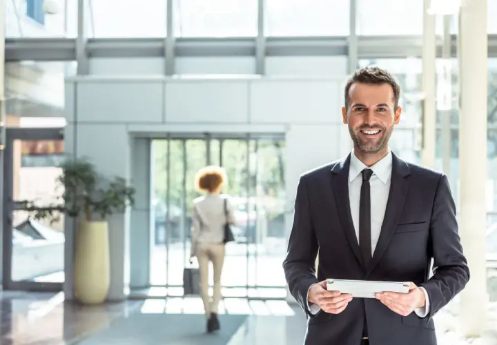 Uomo sorridente in abito scuro con tablet, in un moderno atrio di un pms hotel.