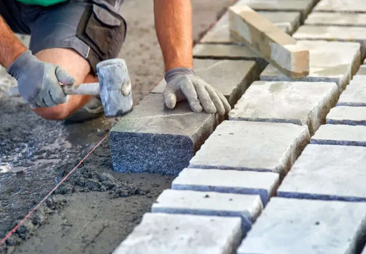 Układanie kostki brukowej na podsypce. Ręce w rękawiczkach kładą kamień, używając gumowego młotka. Widać suchy beton proporcje.