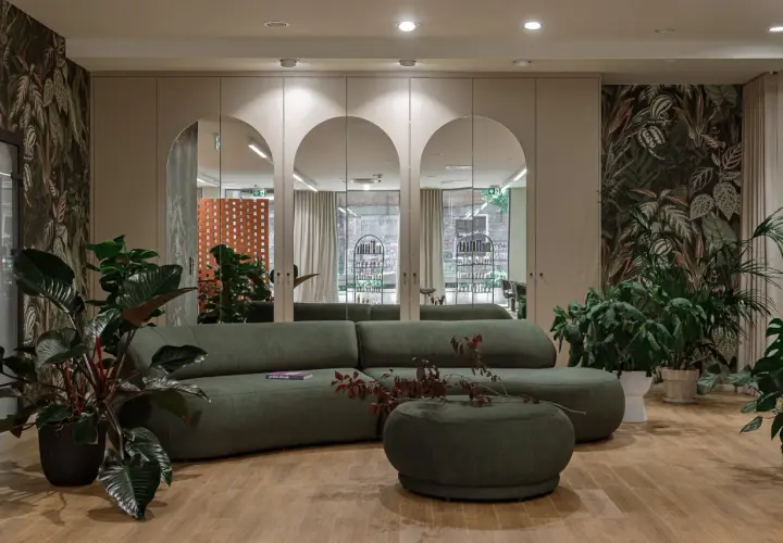 Nowoczesne oświetlenie w salonie fryzjerskim tworzy przytulną atmosferę. Wygodna, zielona sofa i bujna roślinność zapraszają do relaksu.