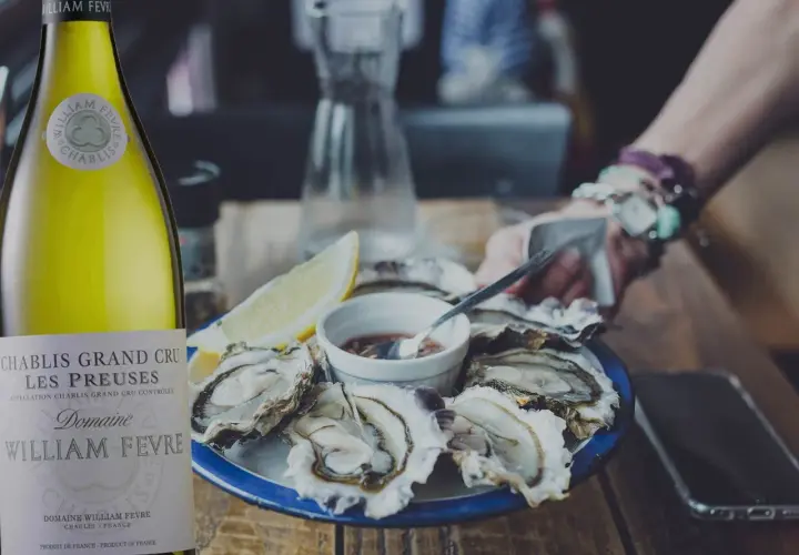 Une bouteille de Chablis Grand Cru William Fèvre, parfaite pour savoir quoi boire avec des huîtres. Une assiette d'huîtres fraîches et un quartier de citron complètent la scène.