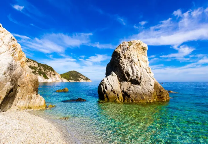 Plage de galets et rochers imposants dans une eau turquoise cristalline, évoquant une charmante ville toscane bord de mer sous un ciel bleu éclatant.