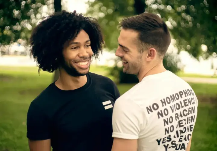 Due uomini sorridono, uno con maglia nera con simbolo di uguaglianza, l'altro con maglia bianca con messaggi di inclusione. Un'immagine che evoca il poliamore significato di amore libero e rispetto.