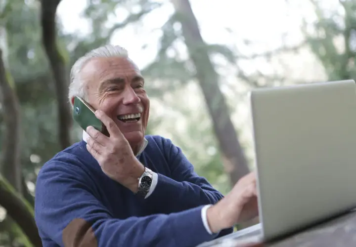 Uśmiechnięty starszy mężczyzna rozmawia przez telefon i pracuje na laptopie na zewnątrz.