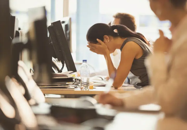 A woman sits at her desk, head in hands, looking demoralized at work. A bottle of water sits nearby.