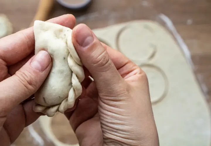 Dłonie formują brzeg pieroga, pokazując, jak lepić pierogi. W tle rozwałkowane ciasto z wyciętymi kółkami.
