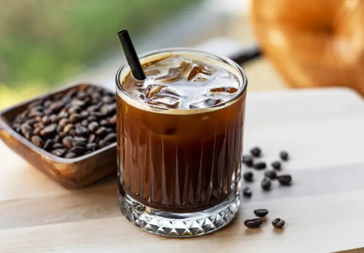 Erfrischender kalter Kaffee mit Eiswürfeln und einem Strohhalm, daneben eine Schale mit Kaffeebohnen.