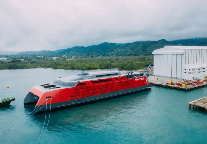 Nowy, czerwono-szary prom Fjord FSTR gotowy do rejsu do Norwegii. W tle zielone wzgórza i biała hala produkcyjna.